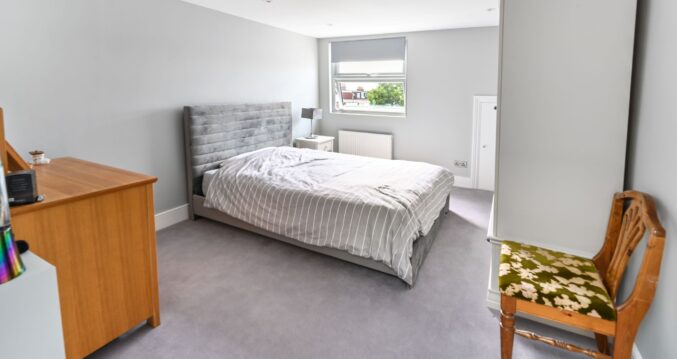 Master bedroom in Barnet loft conversion with dormer window, grey upholstered bed, wooden furniture, and neutral décor