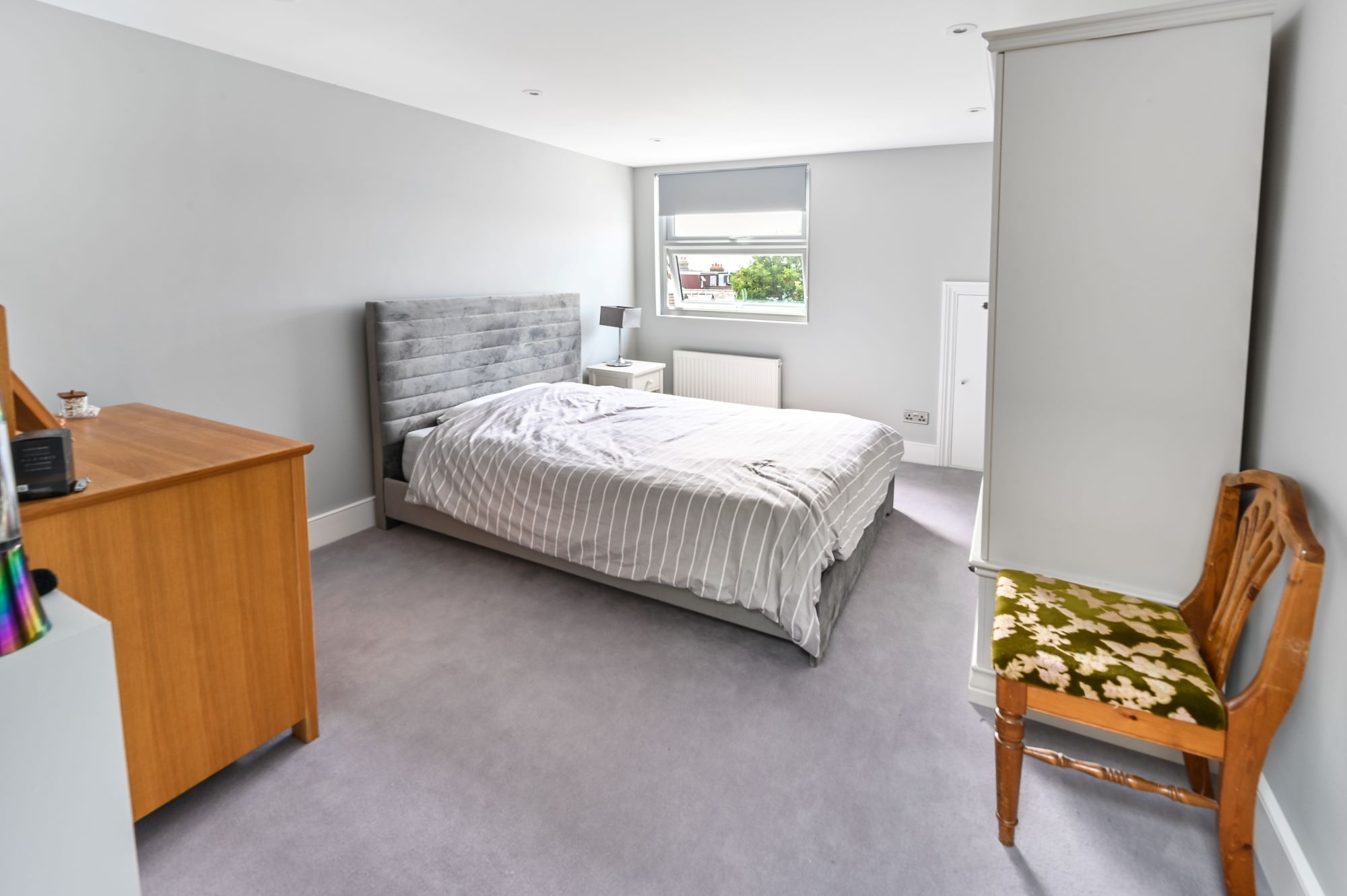 Master bedroom in Barnet loft conversion with dormer window, grey upholstered bed, wooden furniture, and neutral décor