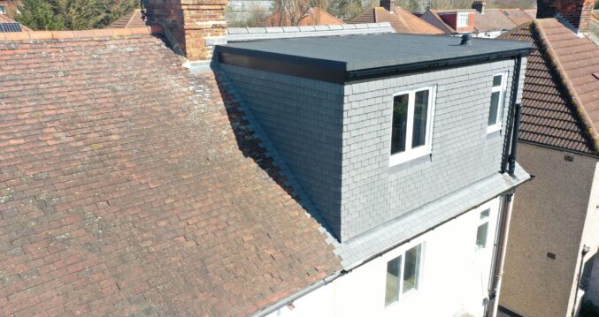 Rear view of a completed hip to gable loft conversion on a Victorian/Edwardian terraced house in Woolwich SE18. The original red tiled roof contrasts with the new grey tiled gable end and flat roof extension, featuring two white uPVC windows and black guttering. Brick chimneys and neighbouring roofs are visible in the background under a clear blue sky.