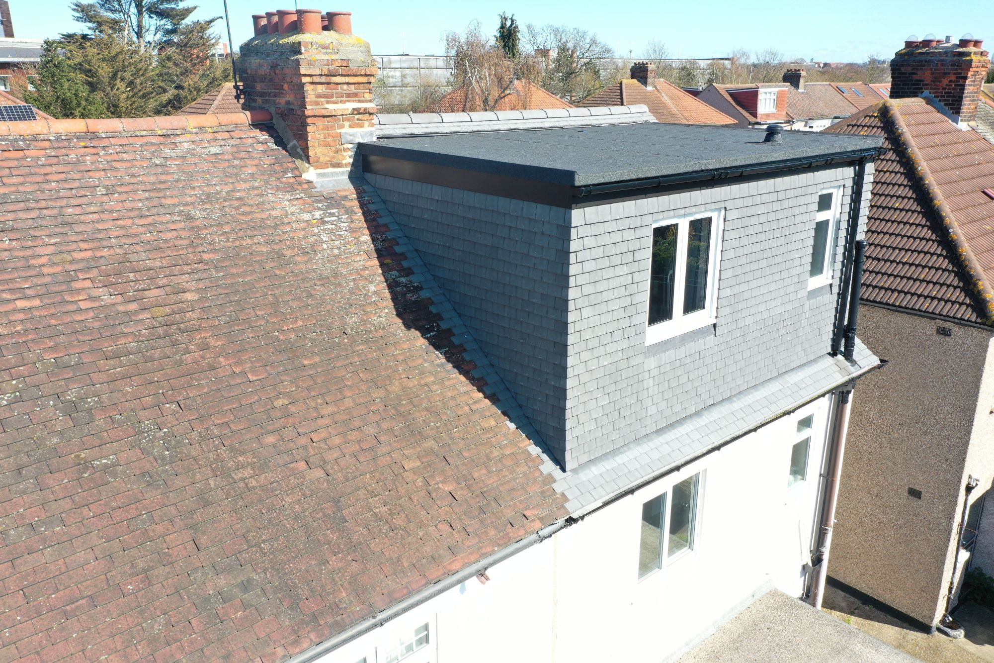 Rear view of a completed hip to gable loft conversion on a Victorian/Edwardian terraced house in Woolwich SE18. The original red tiled roof contrasts with the new grey tiled gable end and flat roof extension, featuring two white uPVC windows and black guttering. Brick chimneys and neighbouring roofs are visible in the background under a clear blue sky.