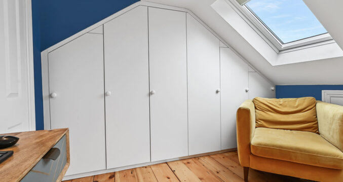 Loft room with fitted wardrobes, blue feature wall, rooflight, timber floor and sofa, supporting Crystal Palace loft storage ideas.