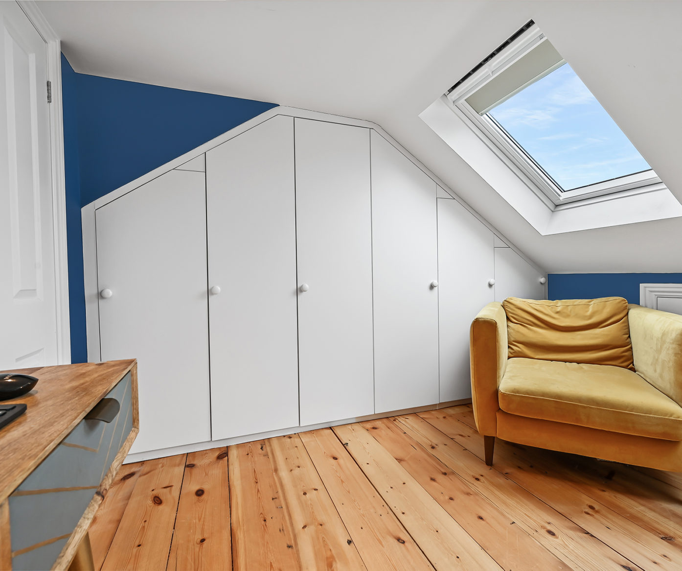 Loft room with fitted wardrobes, blue feature wall, rooflight, timber floor and sofa, supporting Crystal Palace loft storage ideas.